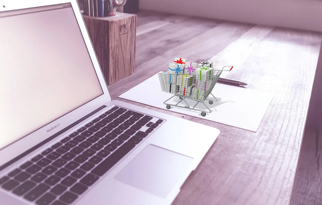Laptop on a desk beside a small shopping cart filled with gift-wrapped boxes, representing online shopping